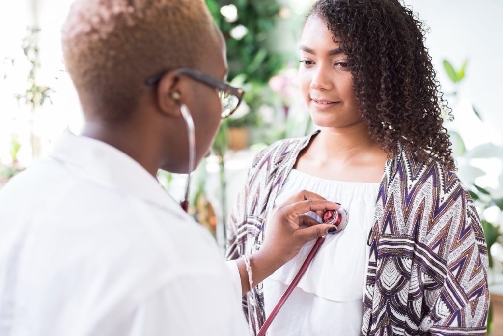 A black woman doctor in a white coat with a stethoscope conducts auscultation of the heart, lungs of a Mexican patient Mixed race young people at the doctors office