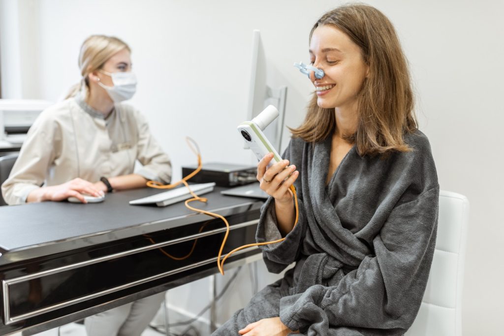 A woman in a grey robe sitting in a medical clinic, holding a handheld FeNO testing device to her mouth while wearing a nose clip during an asthma evaluation.