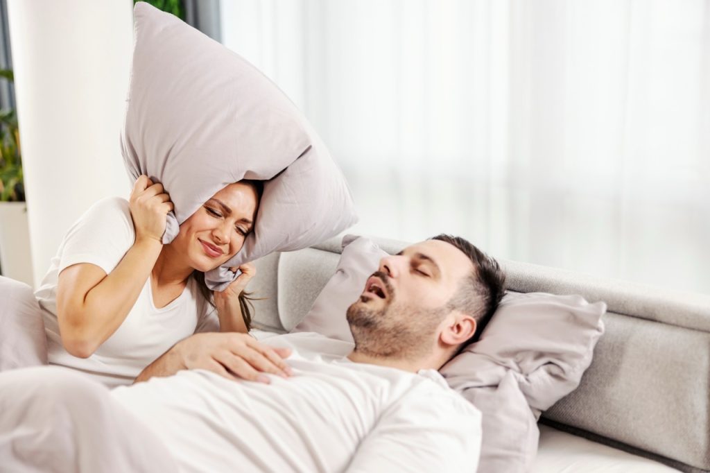 An annoyed woman covering her ears with a pillow in bed while her husband sleeps deeply and snores.