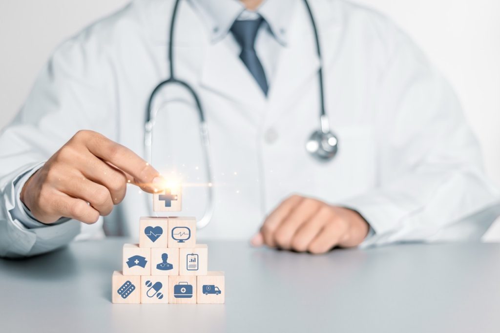 A doctor in a white lab coat sitting at a desk, arranging wooden blocks with healthcare icons including a heart, cross, and stethoscope.