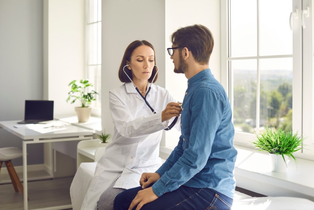 A female physician listening to a male patient's breathing with a stethoscope during a clinic exam.
